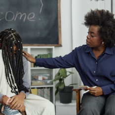 Portrait of caring African American woman comforting teenage girl during therapy session in school and patting on shoulder soothingly 