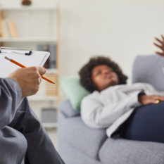 Woman visits professional therapist. Relaxed African American lady lying on sofa and talking about her problems with psychotherapist who is taking notes on paper. Therapy, psychotherapy concept