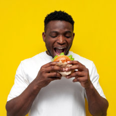 african american man in white t-shirt bites big burger on yellow isolated background, young guy eats fast food 