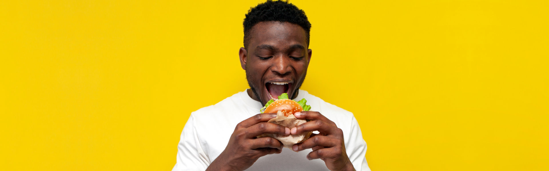 african american man in white t-shirt bites big burger on yellow isolated background, young guy eats fast food